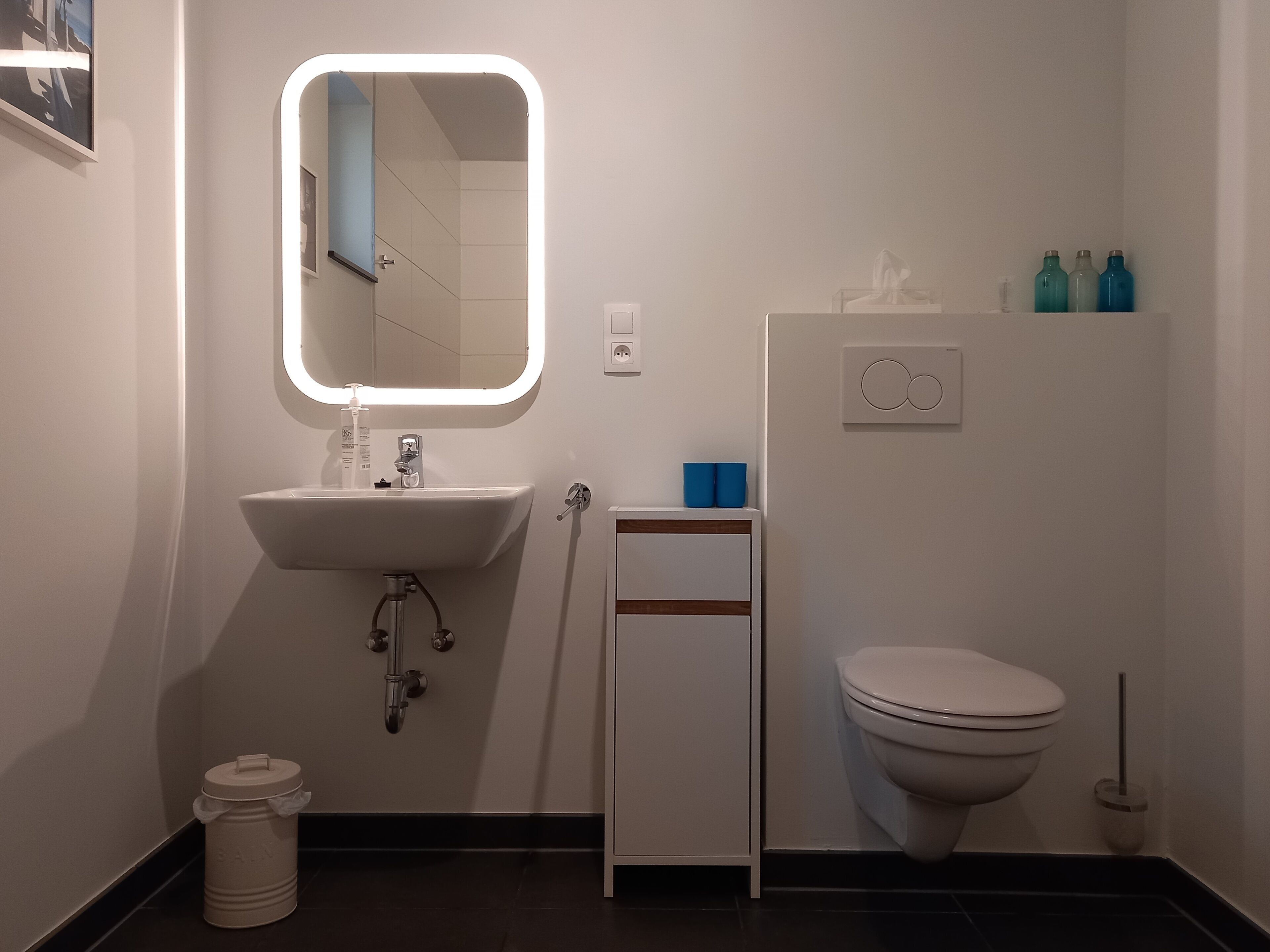 A modern bathroom with a white sink, mirror, and toilet. There are blue bottles on a shelf and a trash bin.
