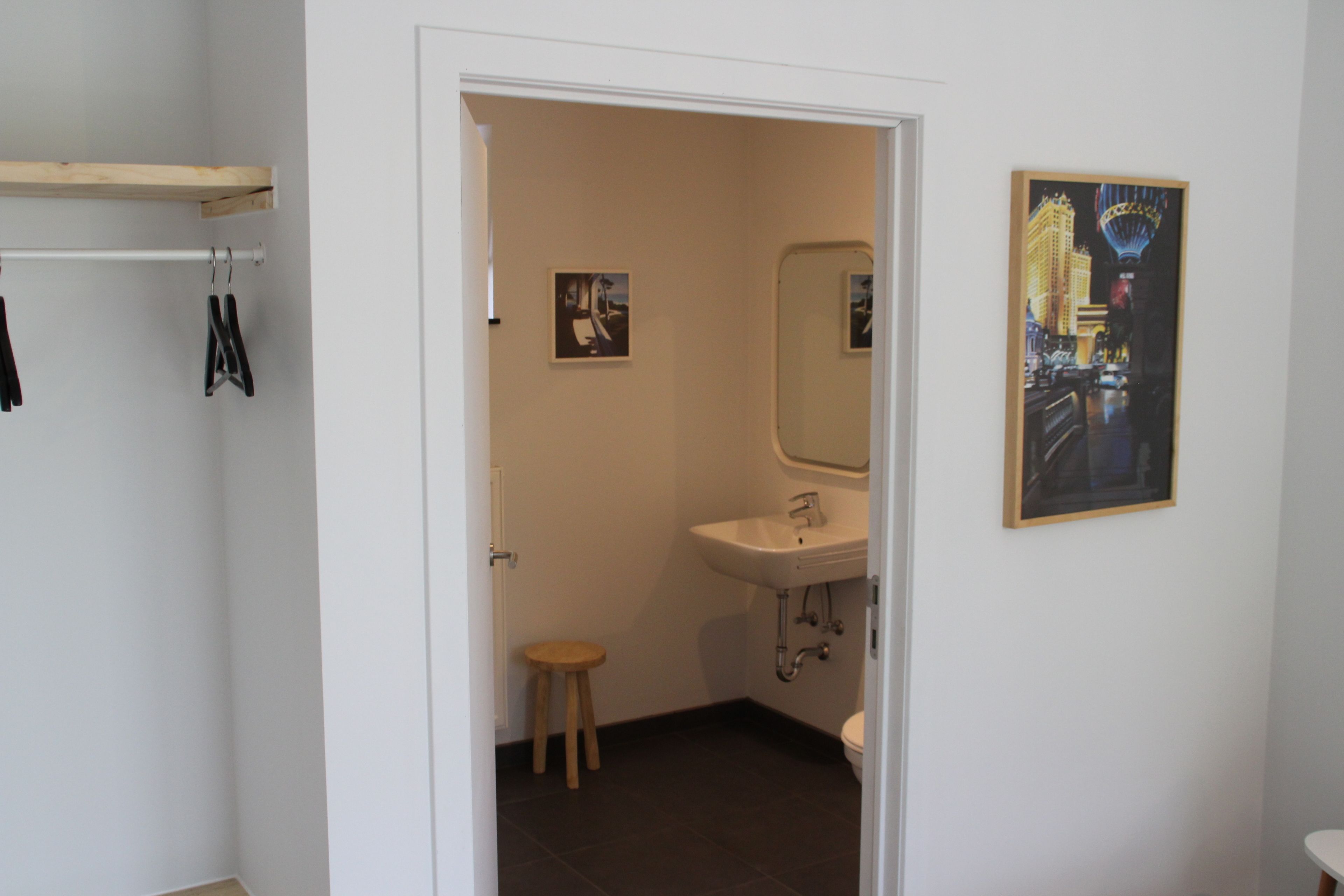Een badkamer met een witte wastafel, een spiegel en een houten krukje. Twee ingelijste foto's hangen aan de muur.