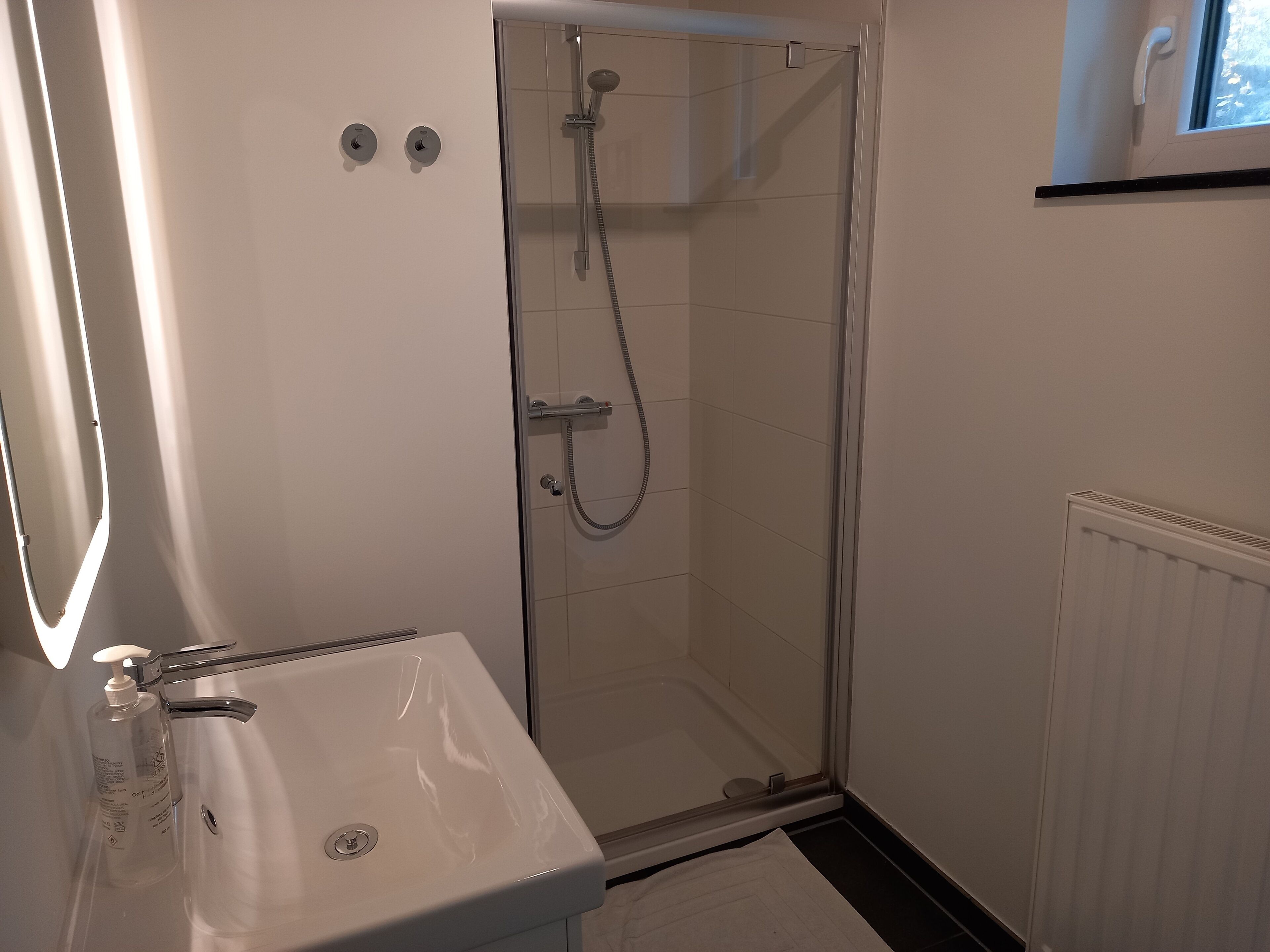 Une salle de bain avec une douche à parois de verre, un lavabo blanc et une fenêtre située au-dessus de la douche.