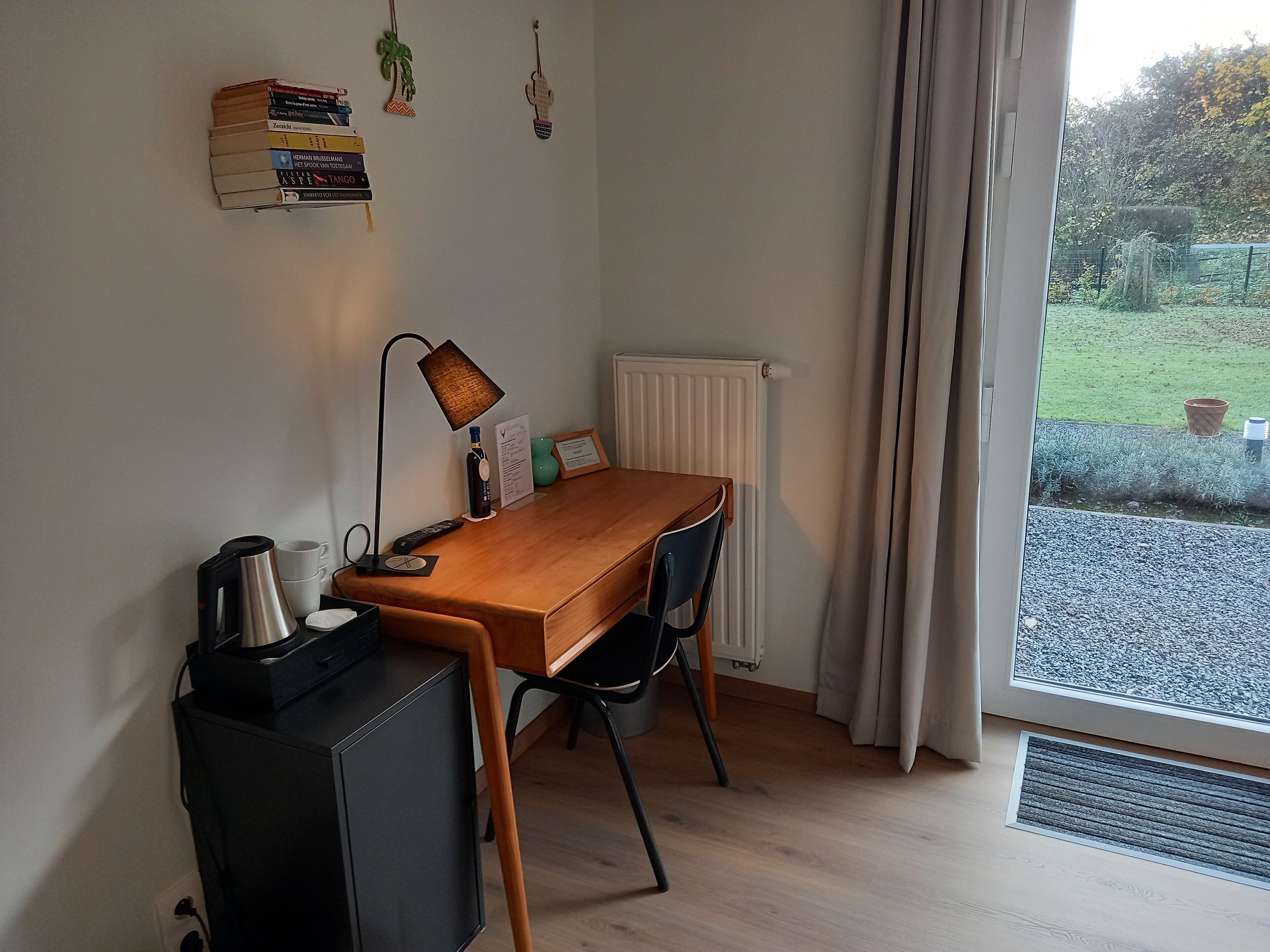 A small, tidy room with a wooden desk, a black cabinet, a lamp, and a radiator. There are books on the wall and a view of a garden through a window.