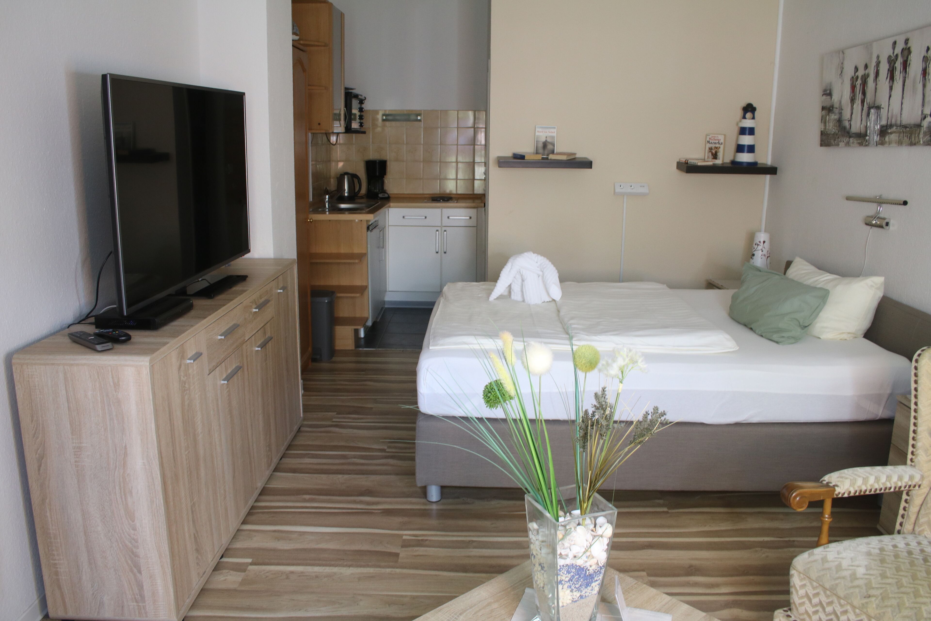 A hotel room with a bed, a TV on a wooden cabinet, a kitchenette, and a dining table with a vase of flowers.