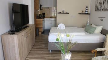A hotel room with a bed, a TV on a wooden cabinet, a kitchenette, and a dining table with a vase of flowers.