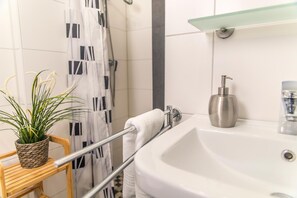 Casa de banho com lavatório branco, prateleira de vidro, cortina de chuveiro, porta-toalhas e uma planta em vaso.