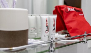 A bathroom counter with RedDoorz shower gel and shampoo, a white cup, and a red bag. - RedDoorz Plus near Paragon Mall Semarang (Semarang)