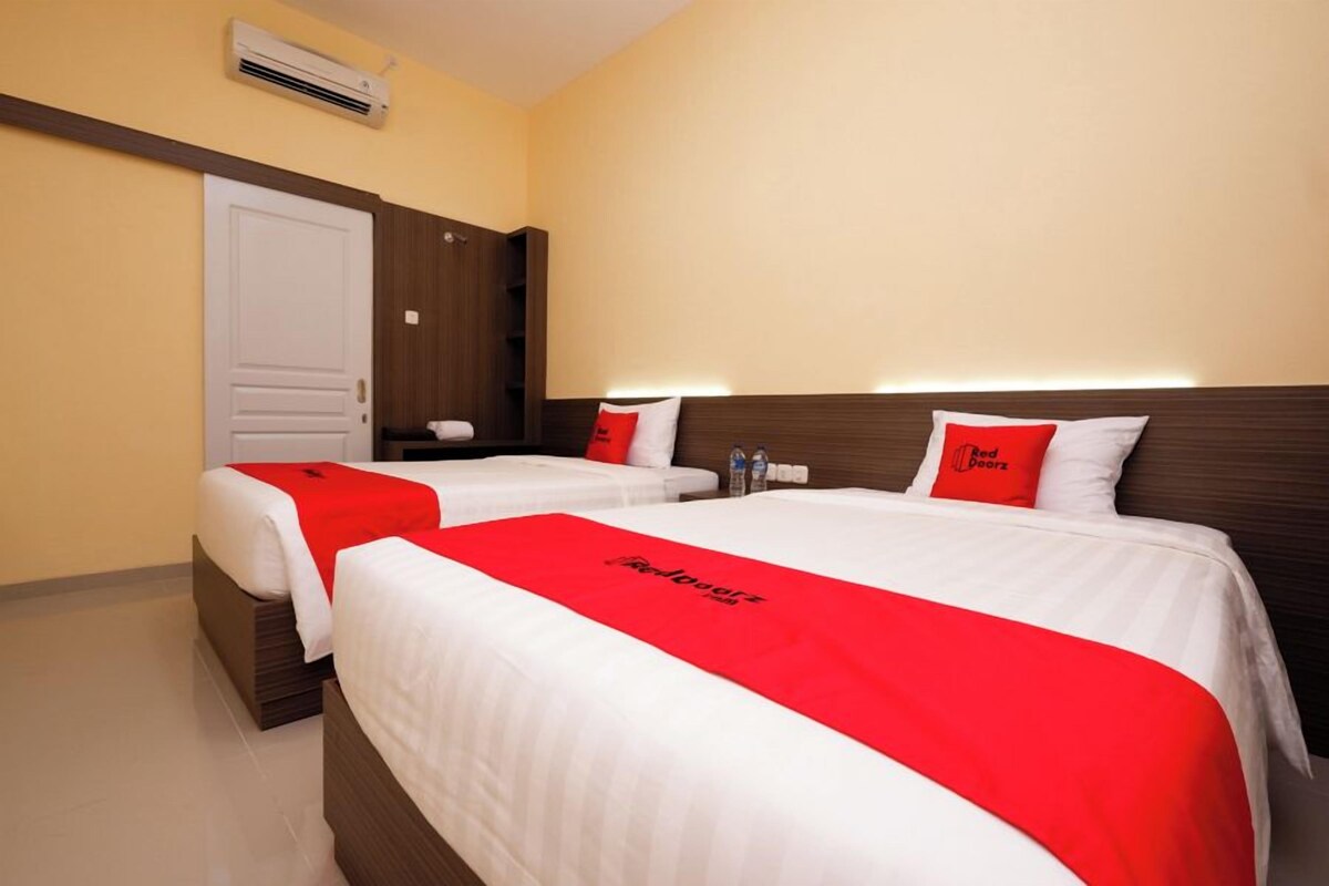 Une chambre d’hôtel avec deux lits, chacun recouvert d’une couverture rouge et agrémenté d’oreillers, une armoire en bois et une porte blanche.