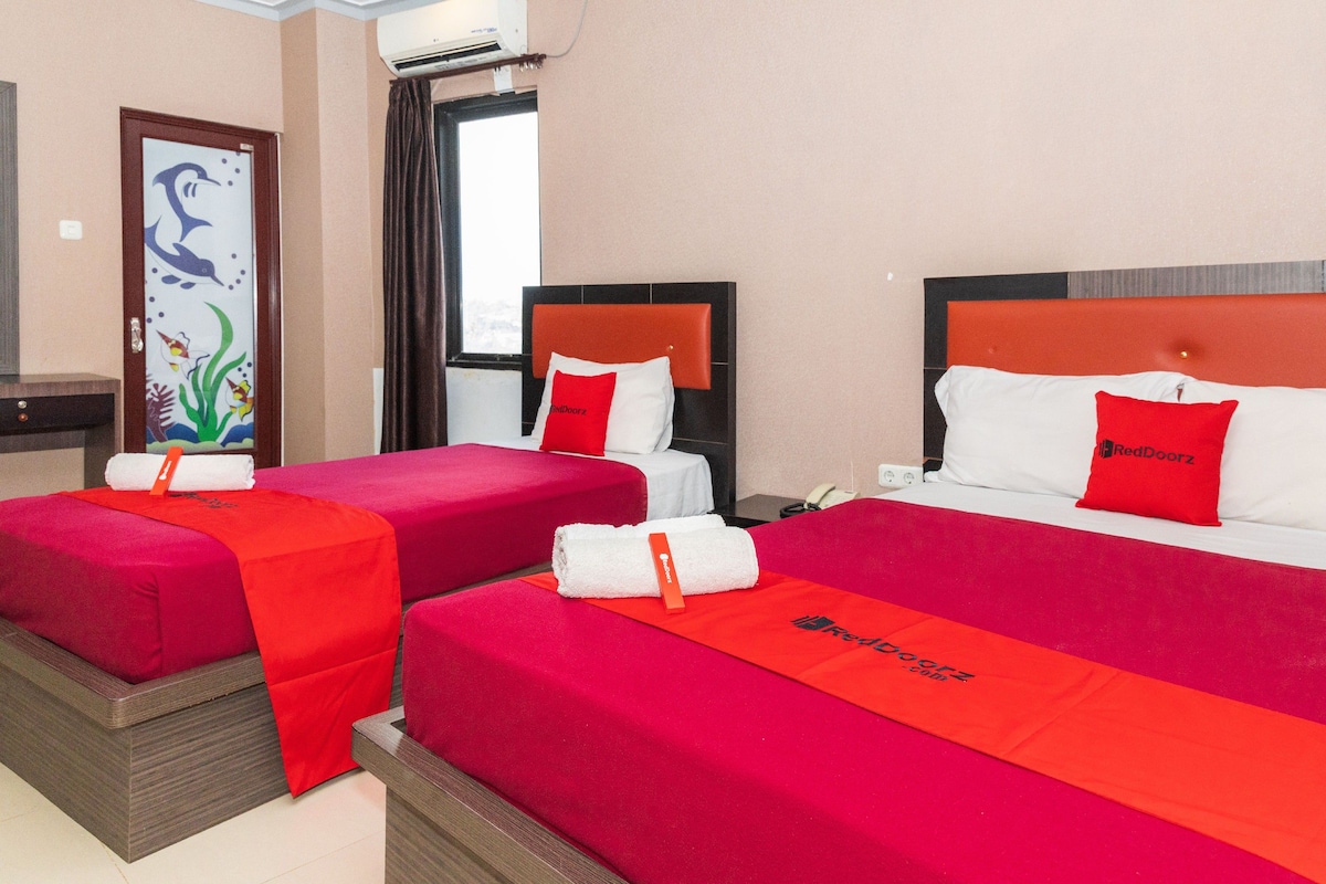 A hotel room with two beds, each with red bedding and a matching headboard. There is a decorative door with a fish and plant design, and a window with curtains.