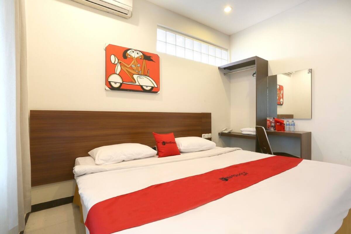 Hotelkamer met een bed, een rode plaid, een houten hoofdbord, een raam, een spiegel en een kunstwerk aan de muur.