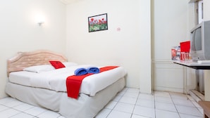 Una cama bien hecha con ropa blanca y detalles rojos, un pequeño cuadro en la pared y un televisor sobre un escritorio.