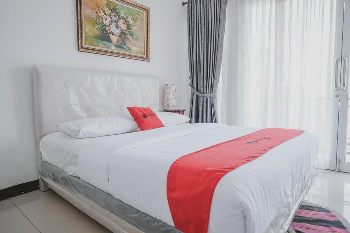 A neatly made bed with white bedding and a red accent pillow, a framed floral artwork above the bed, and a grey curtain to the right.
