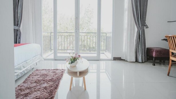 A modern bedroom with a large sliding glass door leading to a balcony, a white bed, a round table with a vase, and a wooden chair.