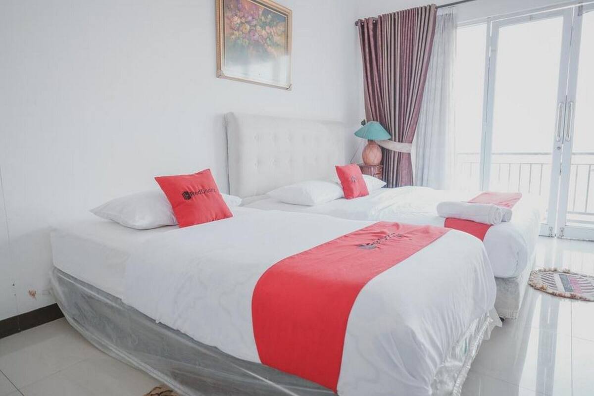 A hotel room with two beds, white bedding, red accents, a lamp, and a view of the outdoors.