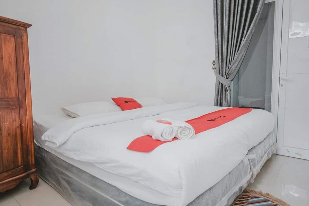 A neatly made bed with white bedding and a red towel, a wooden wardrobe, and a door with a curtain.