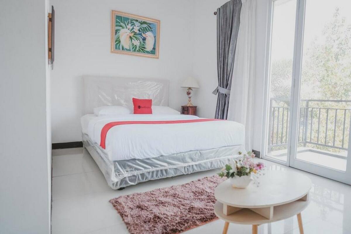 A bedroom with a bed, a small table with flowers, a painting on the wall, and a balcony with curtains.