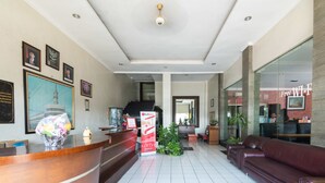 A reception area with a wooden reception desk, framed pictures on the wall, a large potted plant, and a red sofa. - RedDoorz @ Urip Sumoharjo (Surabaya)