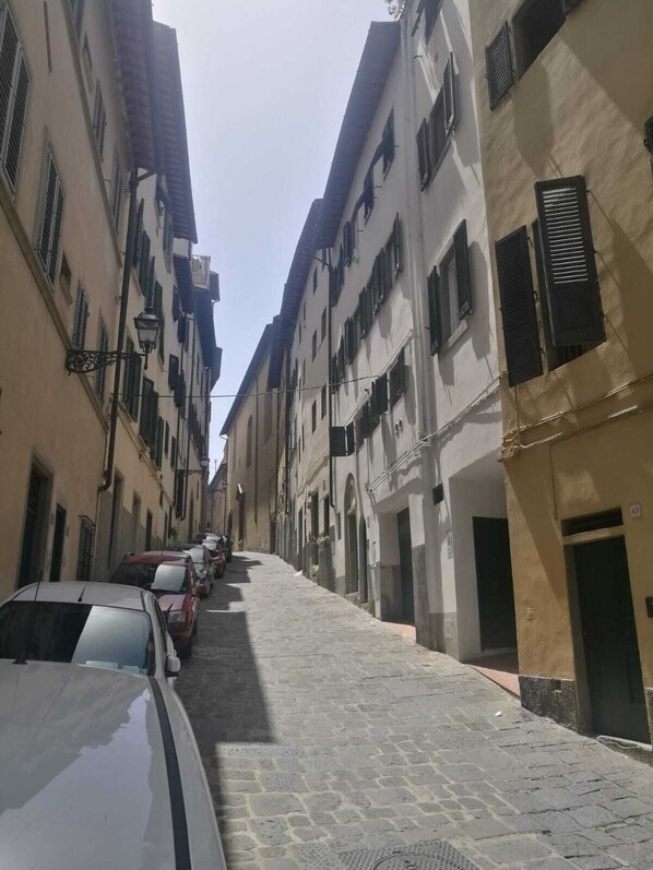 A narrow cobblestone street lined with multi-story buildings, parked cars, and a clear sky.
