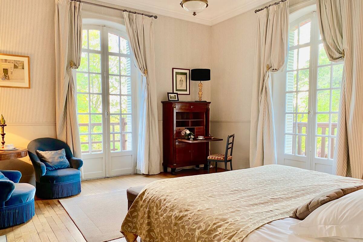 Un dormitorio con una cama grande, dos sillas, un escritorio con lámpara y vistas a la vegetación a través de las ventanas.