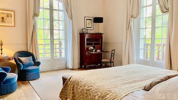 Un dormitorio con una cama grande, dos sillas, un escritorio con lámpara y vistas a la vegetación a través de las ventanas.
