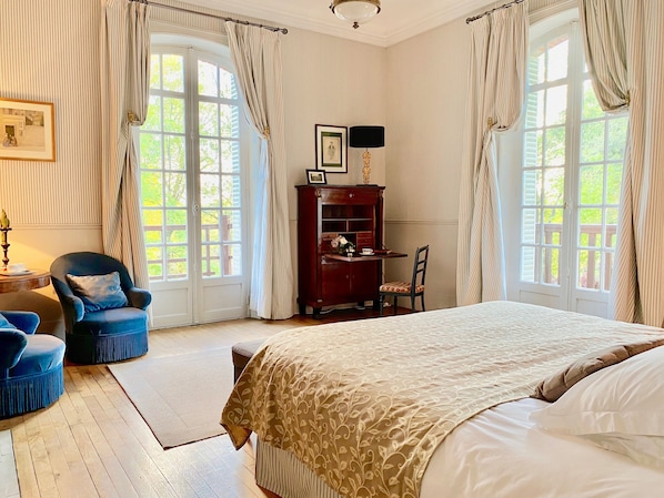 Ein Schlafzimmer mit einem großen Bett, zwei Stühlen, einem Schreibtisch mit Lampe und Blick auf Grün hinter den Fenstern.