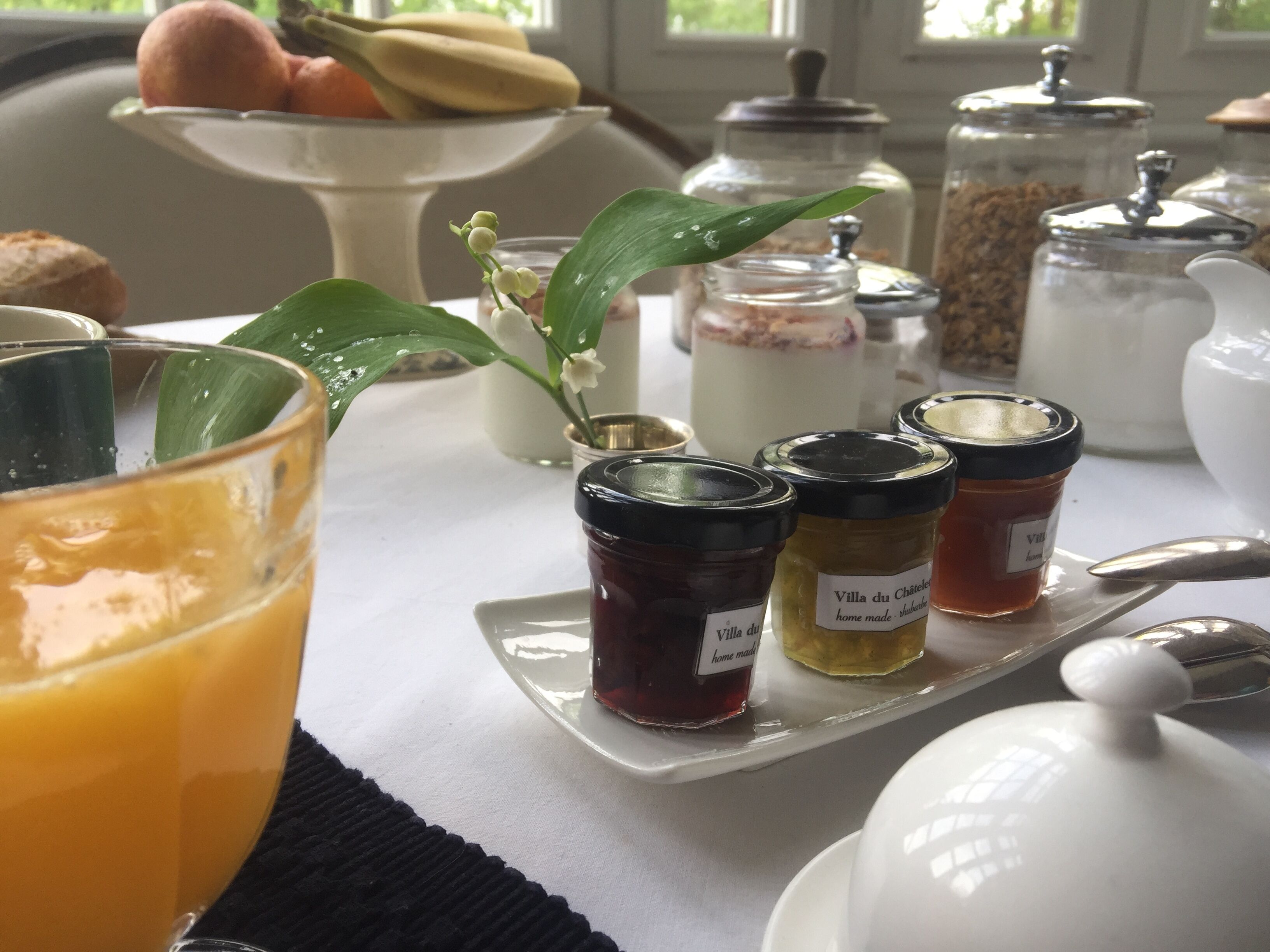 Desayuno con un vaso de jugo de naranja, una tetera blanca y frascos de mermelada sobre una bandeja.