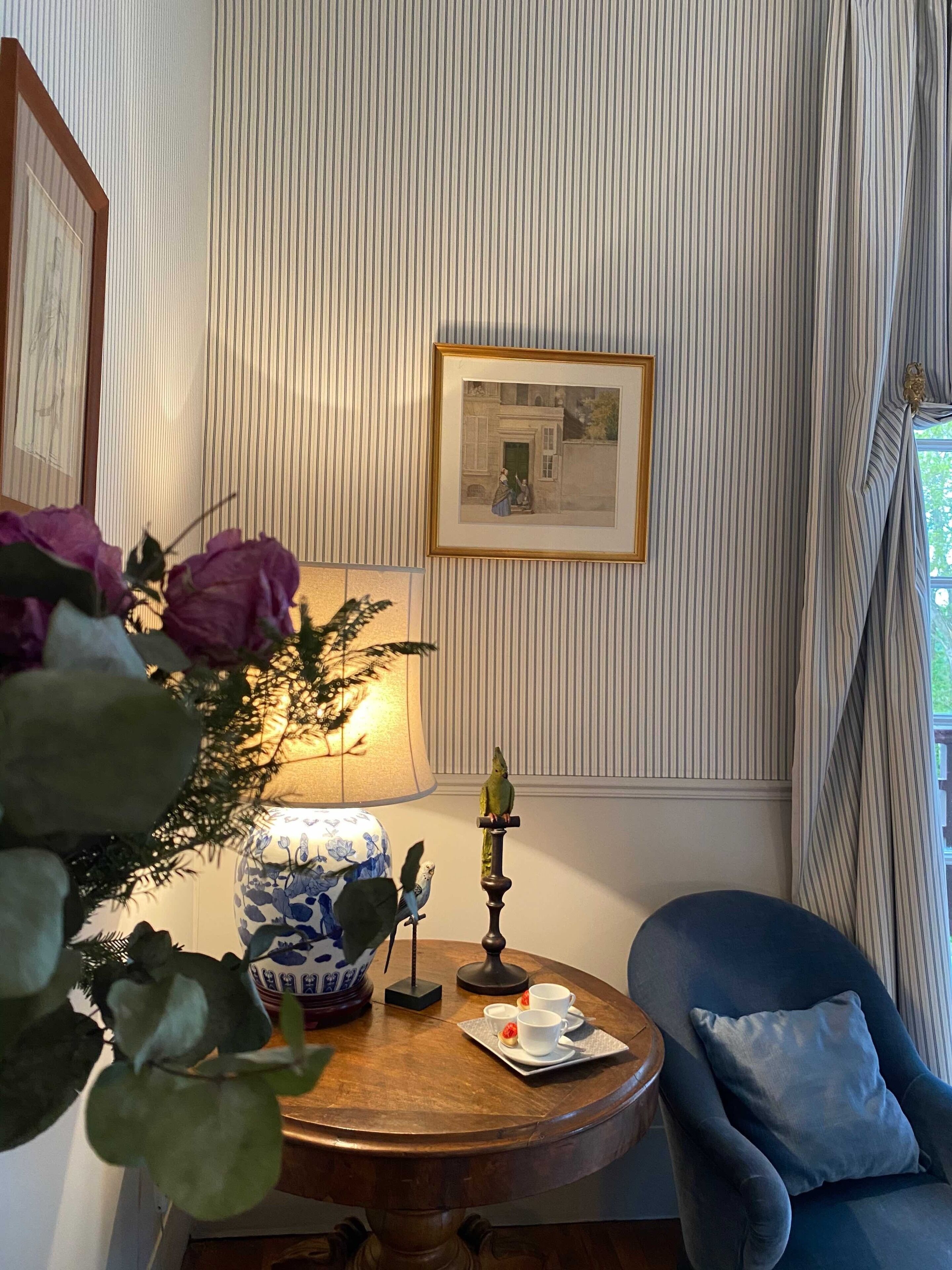 A room with striped wallpaper, a framed picture, a table with a lamp, a vase, and a chair.