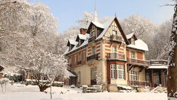 Un edificio grande e histórico con varios balcones y una torre destacada, rodeado de árboles cubiertos de nieve.