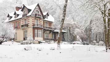 A large, historic building with multiple stories and a prominent balcony, surrounded by snow-covered trees and a fence.