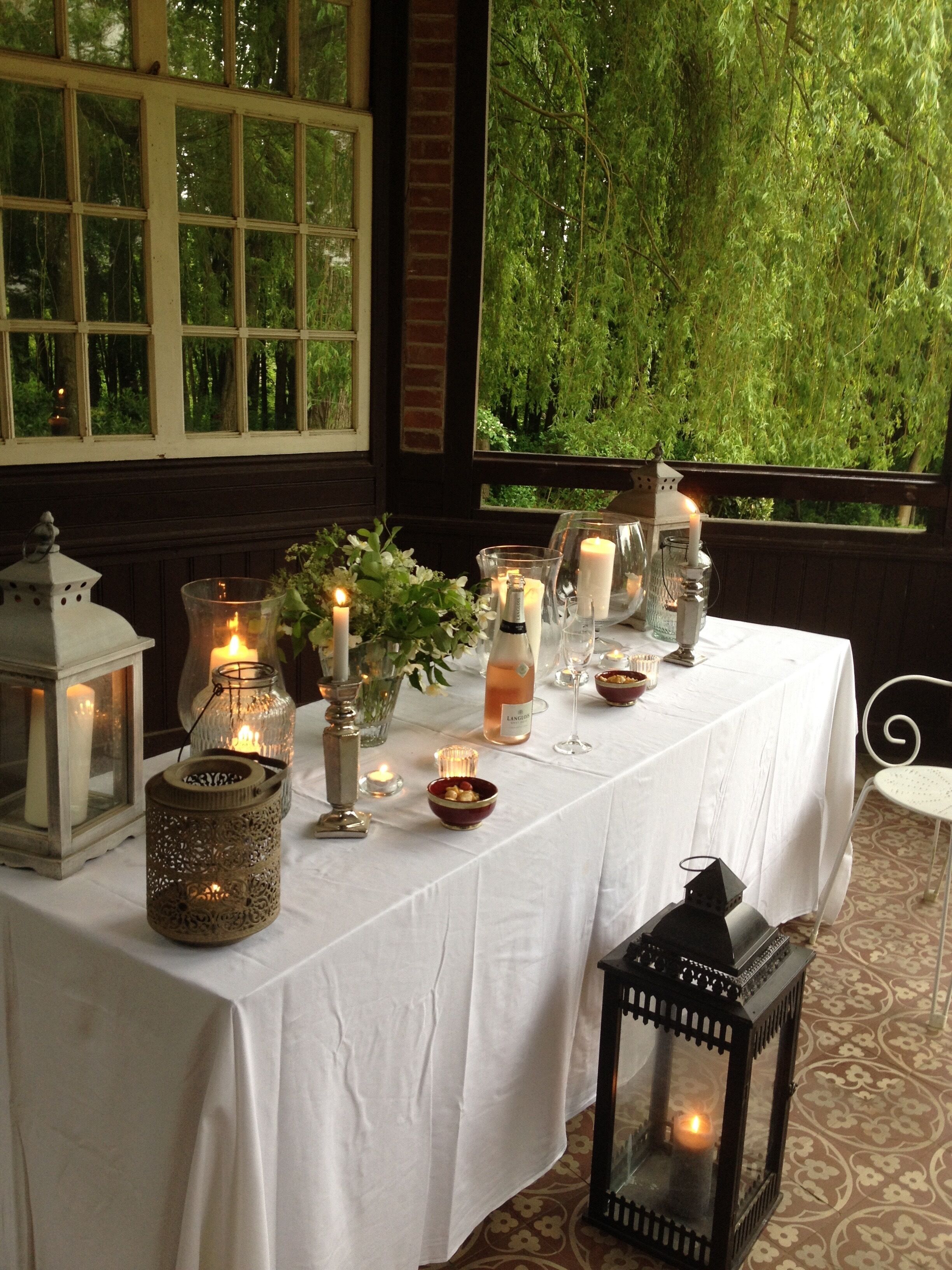 Mesa servida con velas, cristalería y una botella de vino, con vista a follaje a través de una ventana.