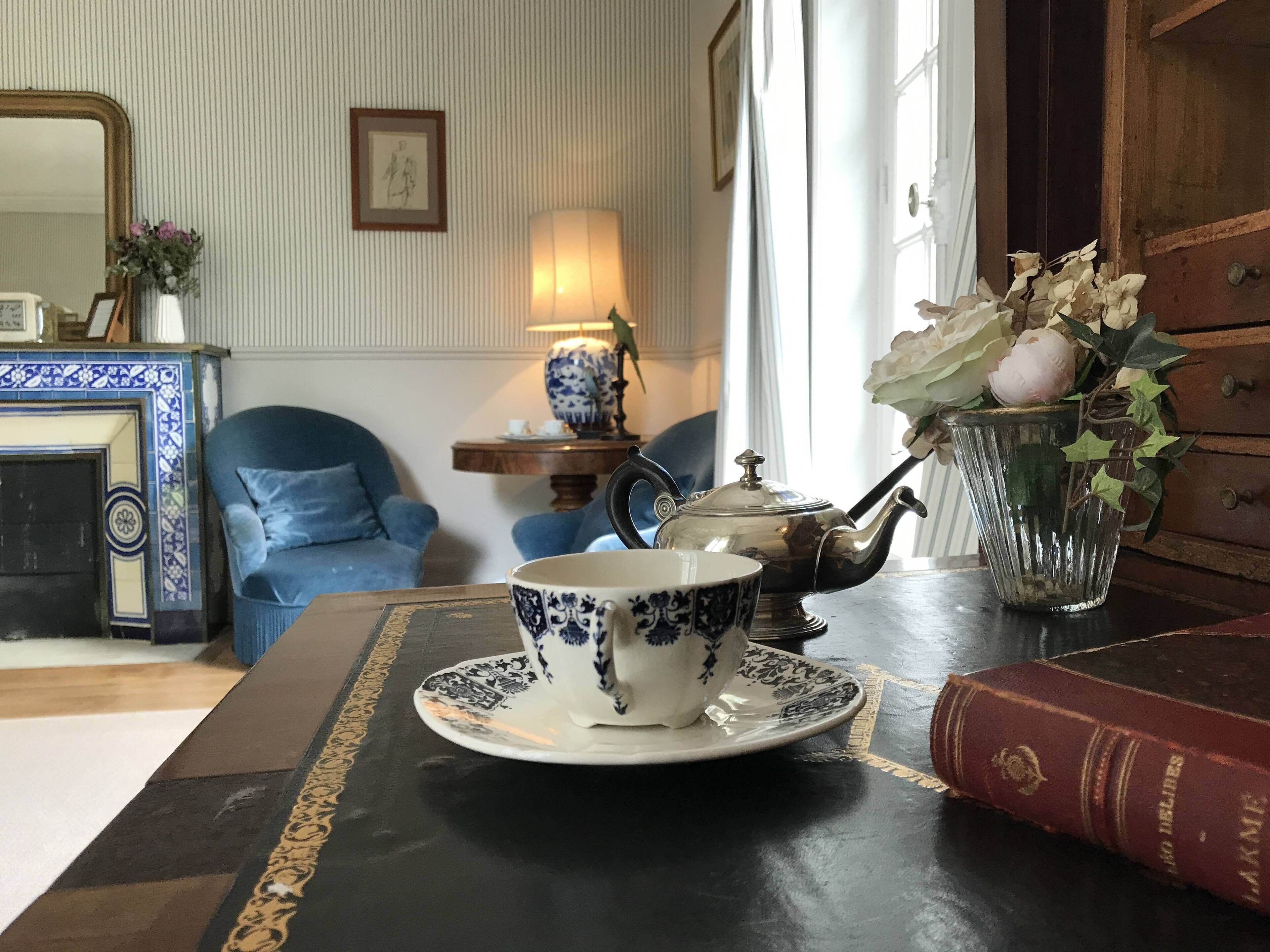 Una habitación con una mesa preparada para el té, un sillón azul, un aparador de madera y una estantería con un libro.
