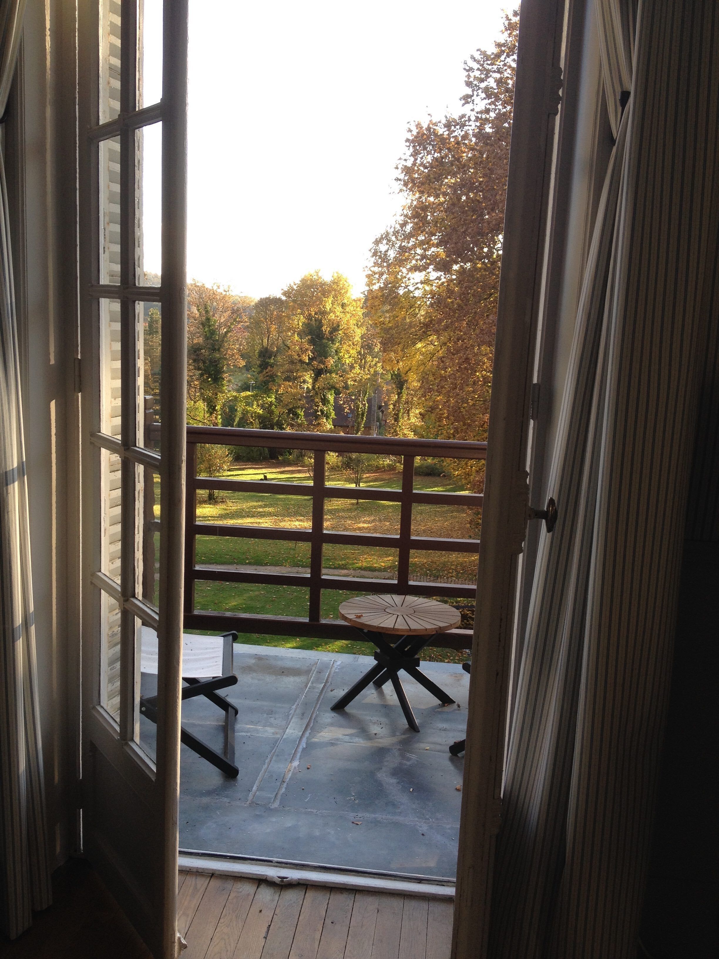 Une porte ouverte donnant sur un balcon avec une table et des chaises, une vue sur une zone herbeuse et des arbres, et un bâtiment en arrière-plan.