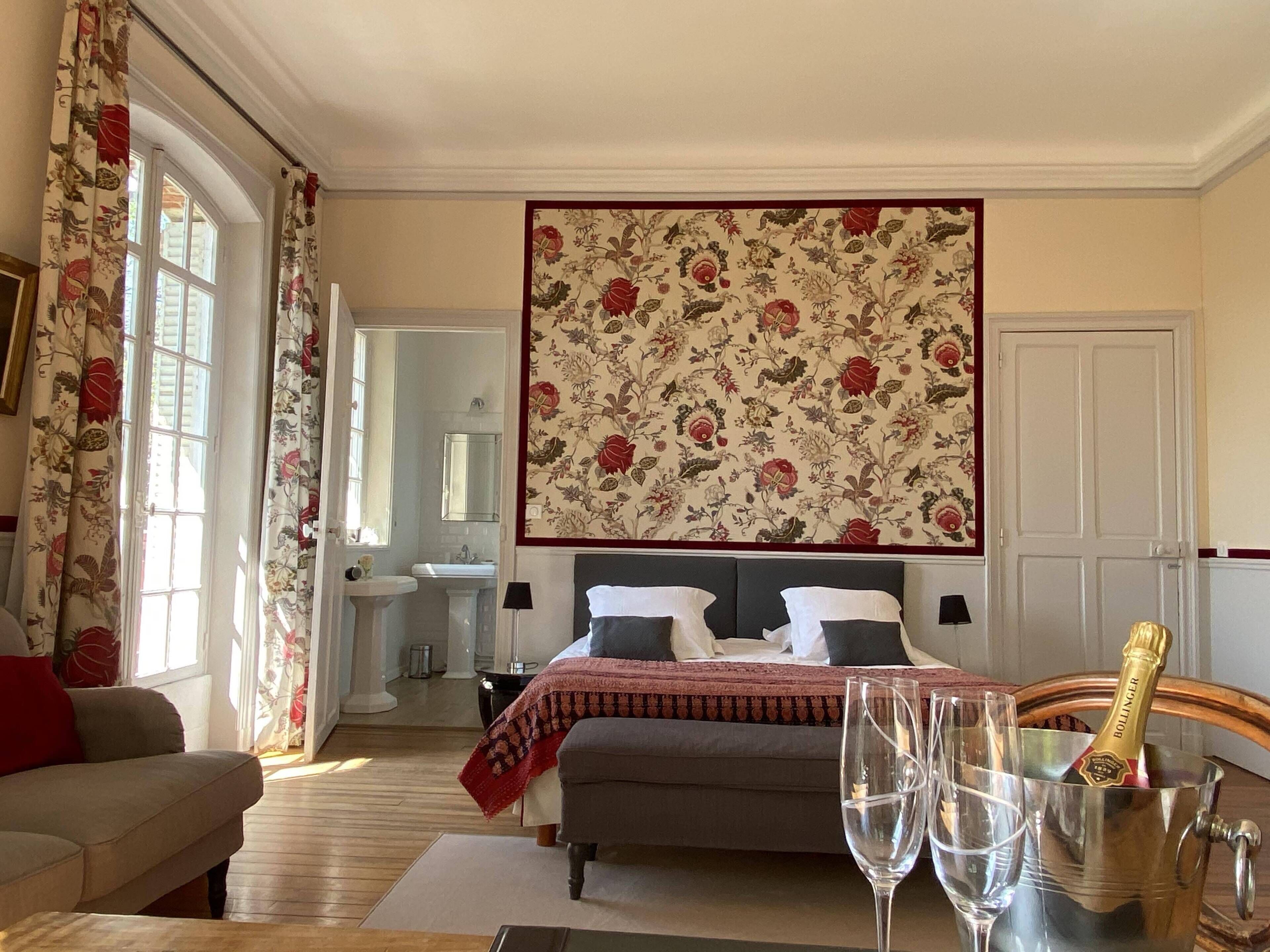 A bedroom with a bed, bedside tables, a nightstand, a window with curtains, a bathroom with a sink and mirror, and a large floral-patterned wall hanging.