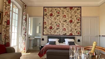 A bedroom with a bed, bedside tables, a nightstand, a window with curtains, a bathroom with a sink and mirror, and a large floral-patterned wall hanging.