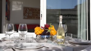 Una mesa de comedor dispuesta con platos, vasos y cubiertos, una botella de vino blanco, y un cuenco con pimientos amarillos y berenjena.