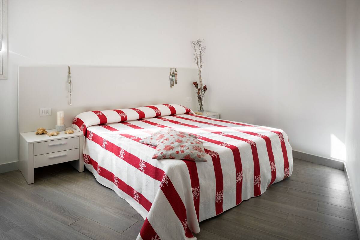 Un lit bien fait, recouvert d’une couvre-lit à rayures rouges et blanches, une table de chevet avec une lampe et un vase contenant des fleurs séchées sur la table de chevet.