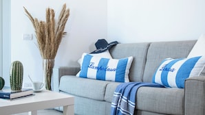 Una sala de estar con un sofá gris, almohadas a rayas, una manta azul y blanca, un cactus, una taza blanca y un libro sobre una mesa blanca.
