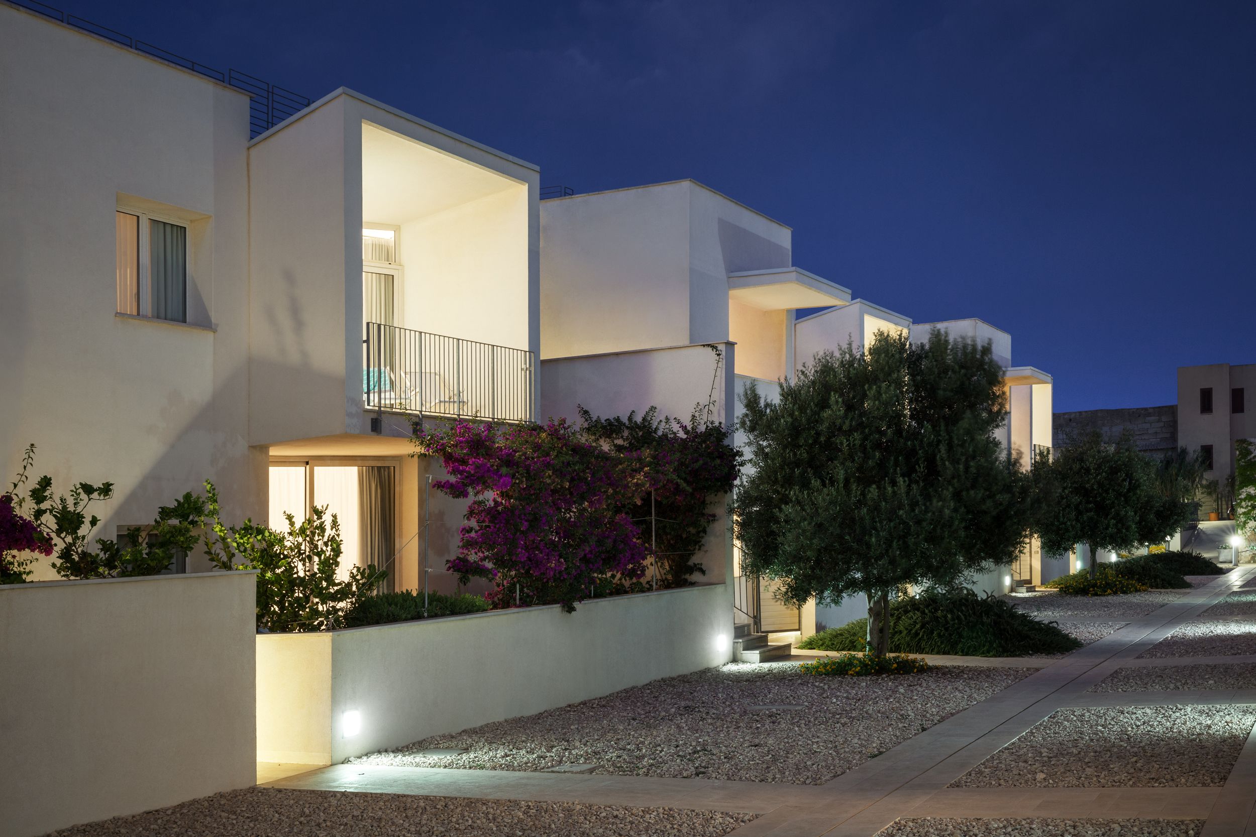 Un immeuble résidentiel moderne doté d’un balcon, d’un hall d’entrée bien éclairé et d’un chemin recouvert de gravier.
