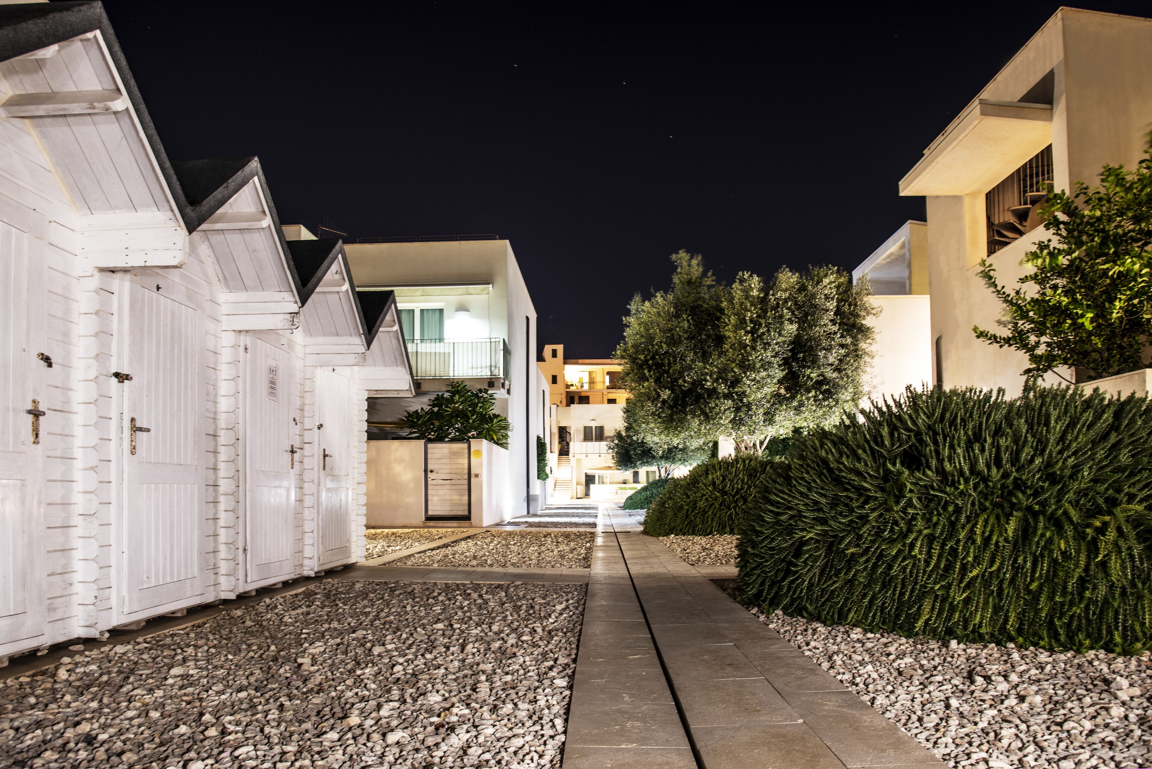 A nighttime view of a residential street with houses, a gravel pathway, and a hedge.