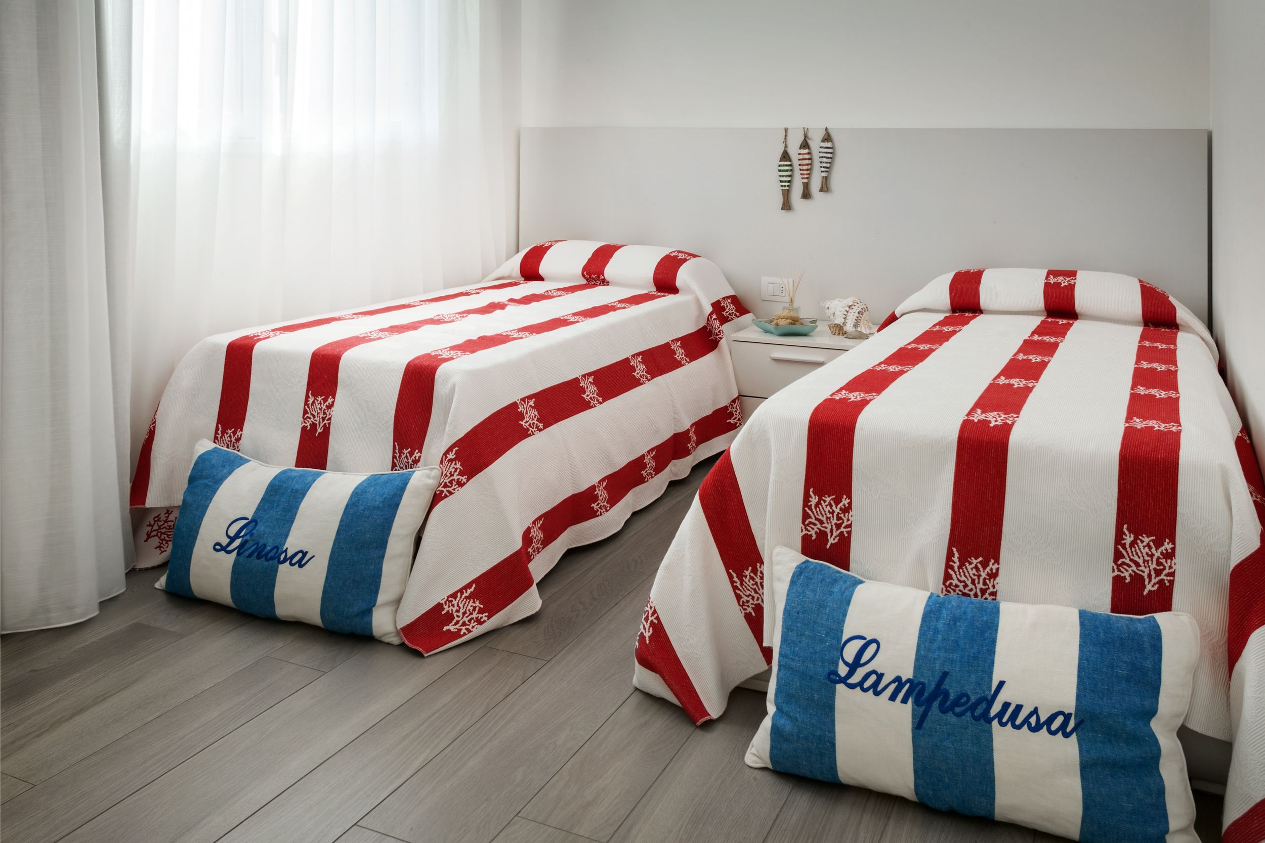 A hotel room with two beds, each with red and white striped bedding and pillows labeled "Lima" and "Lampedusa."