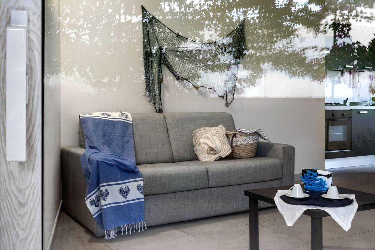 Un salon avec un canapé gris, une couverture bleue et blanche, un filet décoratif et une petite table recouverte d’une nappe blanche.
