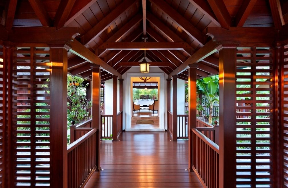 A wooden covered walkway with a central table and chairs, flanked by wooden railings and greenery.