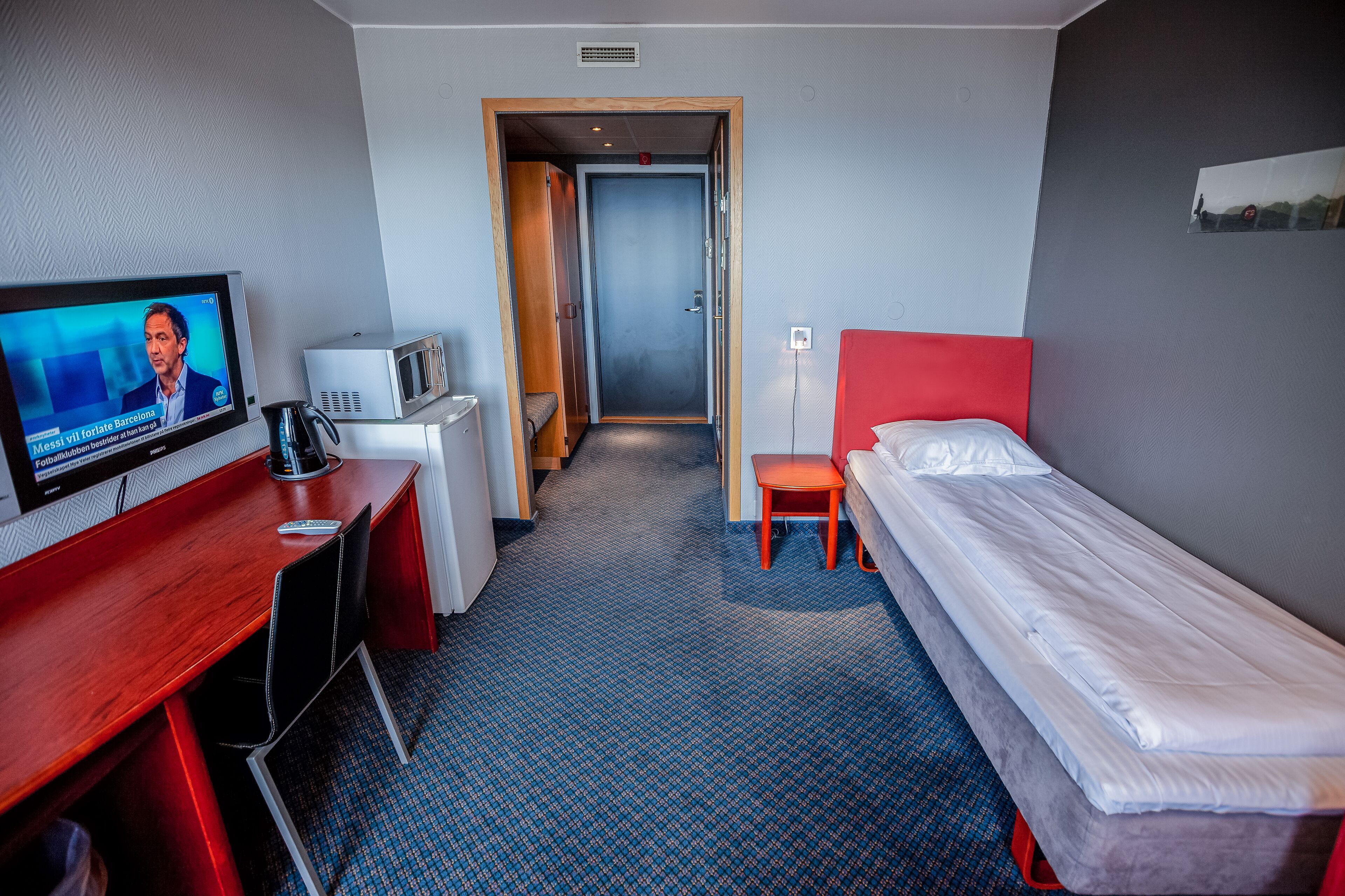 Et hotelværelse med seng, skrivebord med fjernsyn, mikroovn og en rød stol.