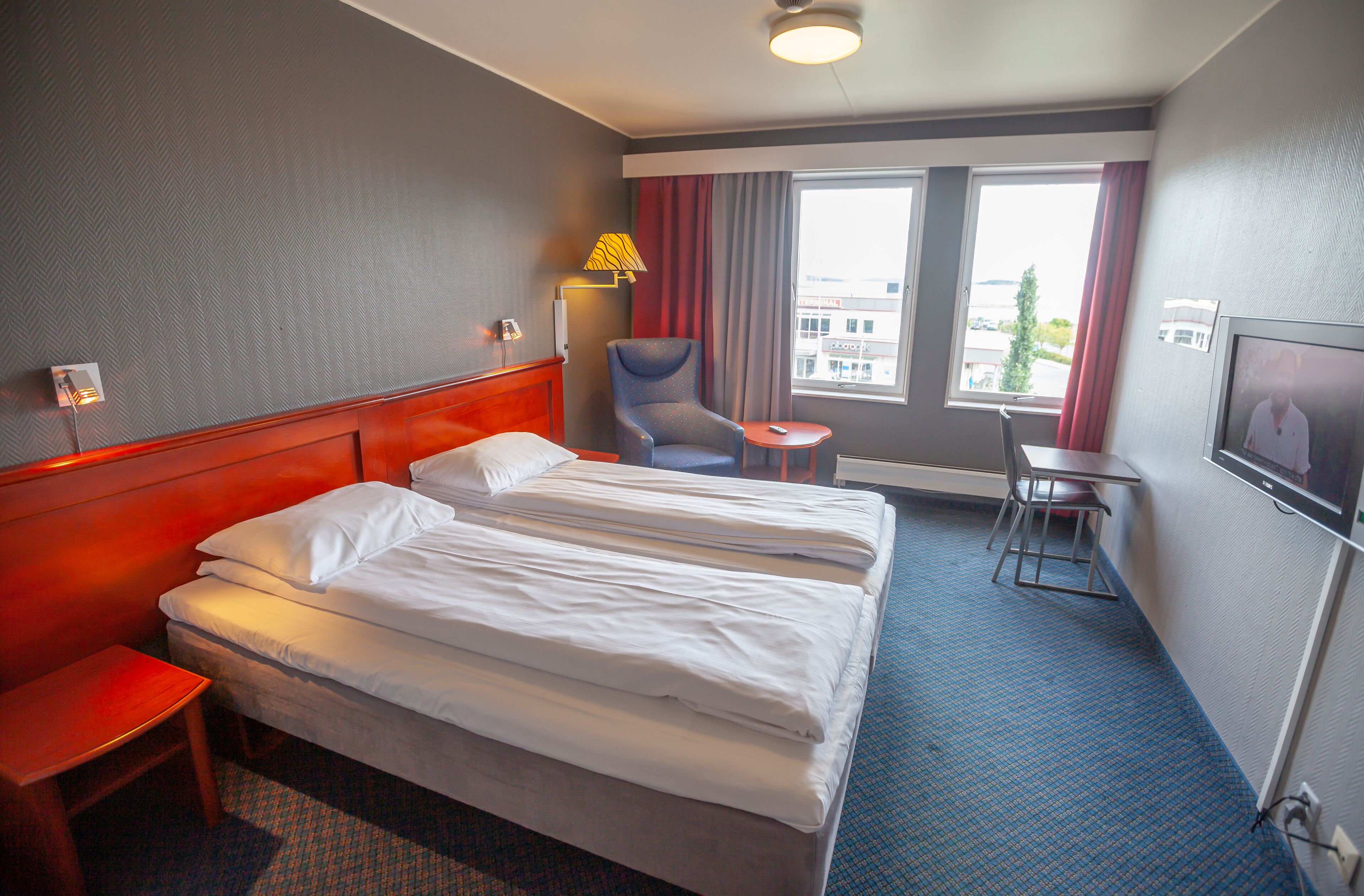 A hotel room with a double bed, a chair, a desk, and a window with curtains.