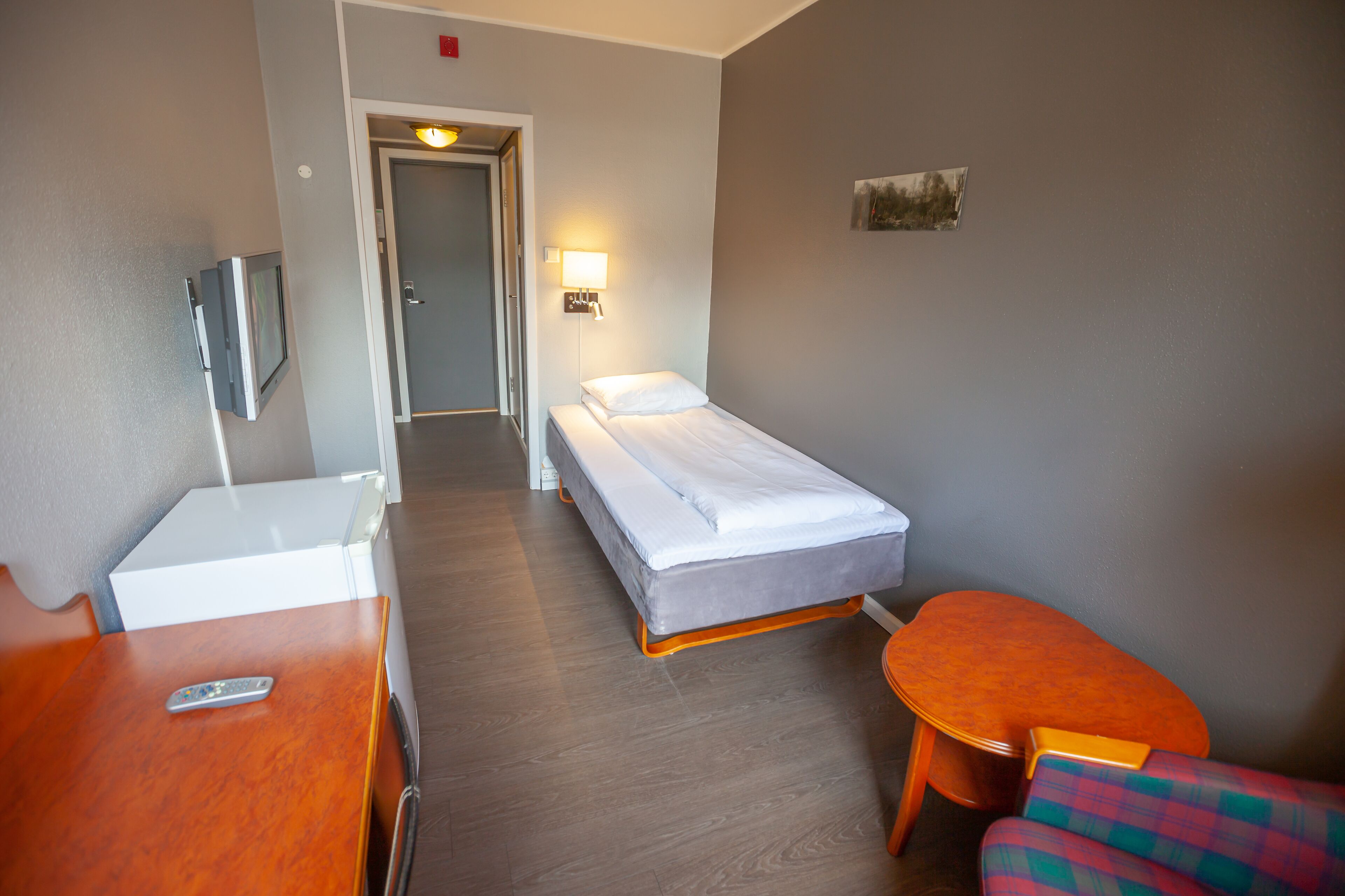 Habitación de hotel con una cama individual, una mesita, una silla, un televisor y una lámpara.