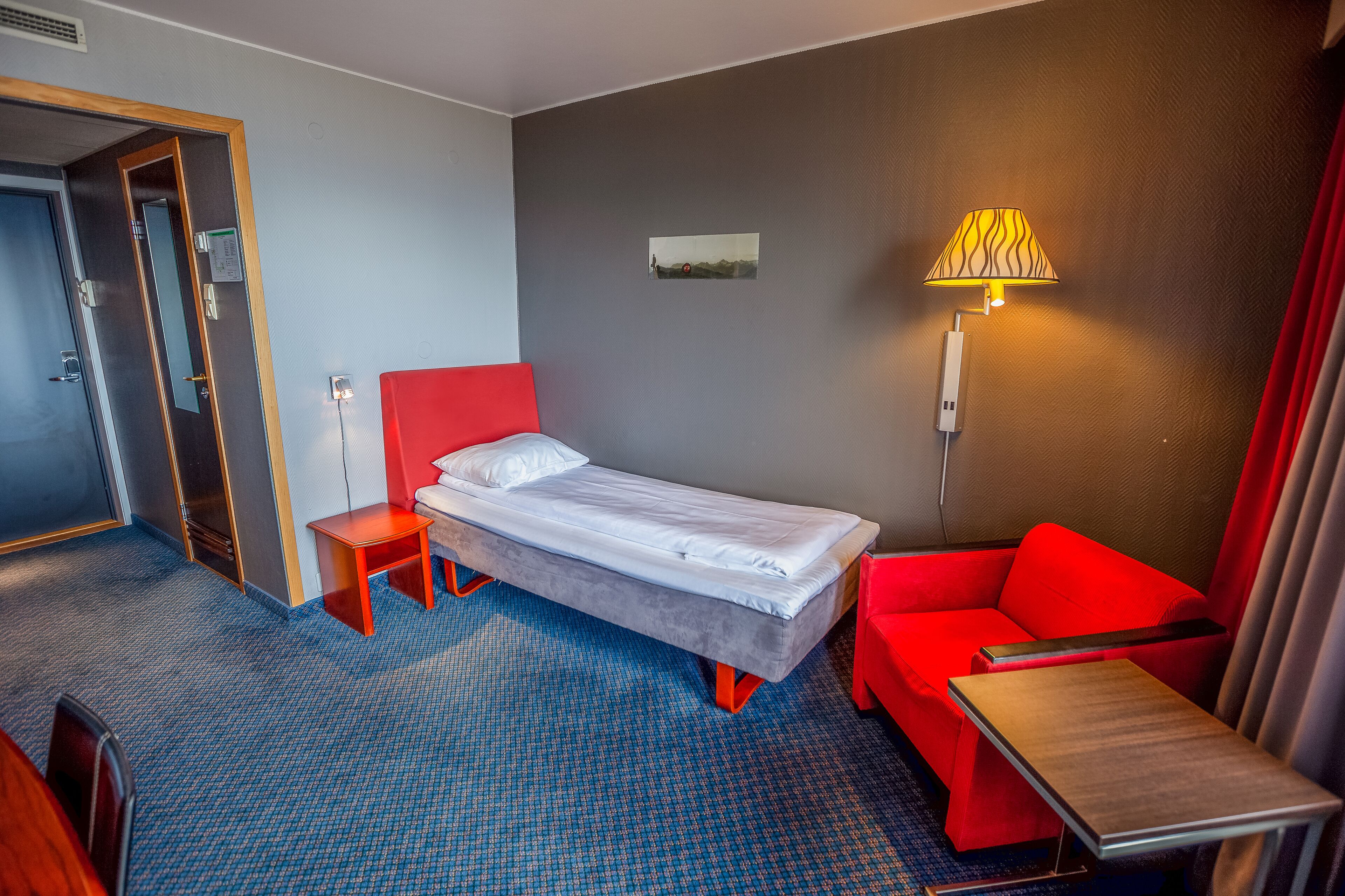 A hotel room with a single bed, a red sofa, a small table, and a lamp.