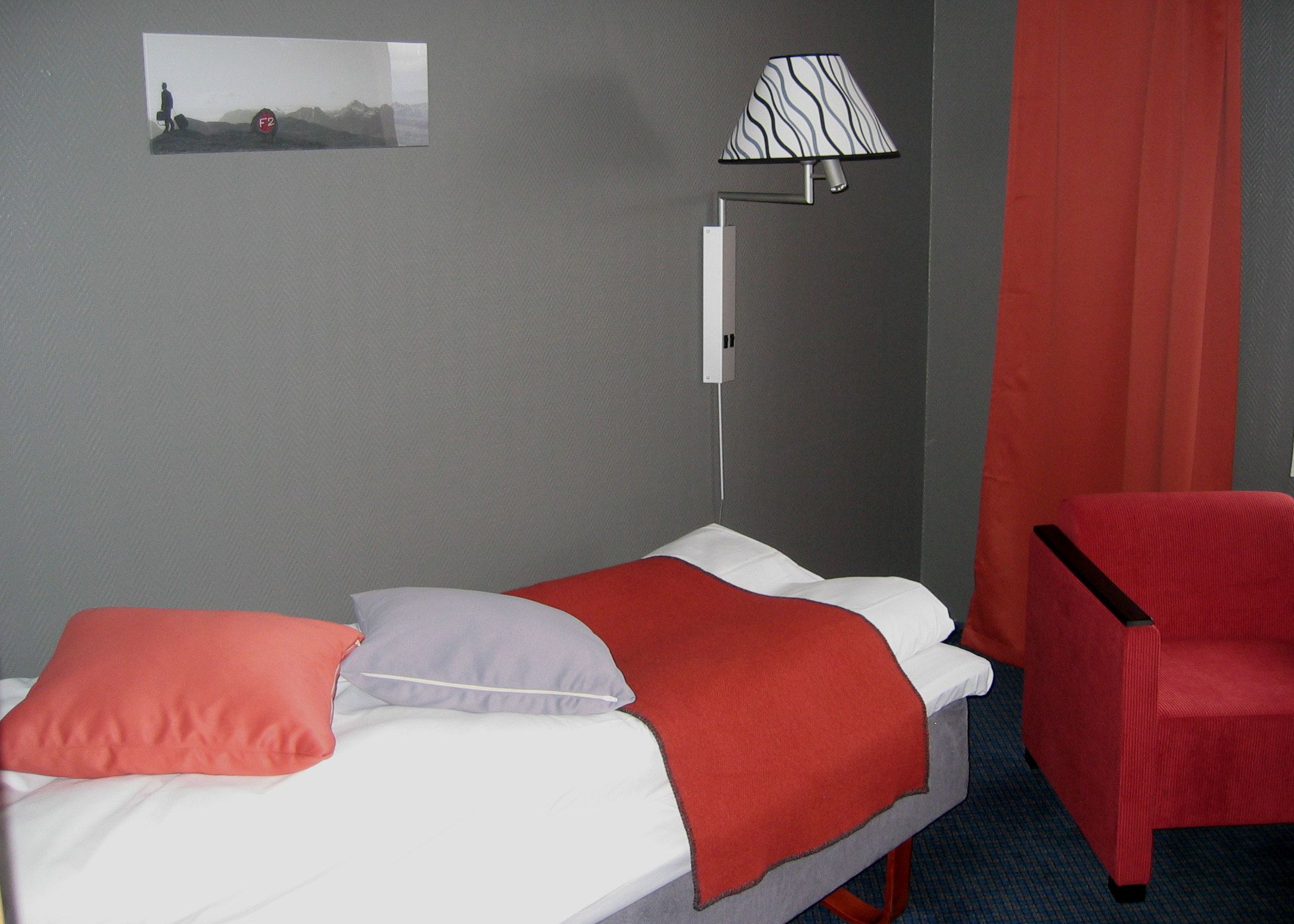 Habitación de hotel con una cama, una silla roja, una lámpara y un cuadro en la pared.