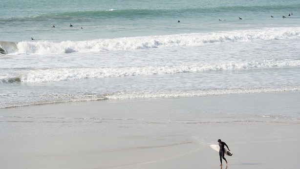 A beach with waves, a surfer walking on the sand, and several surfers in the water.