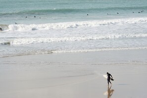 Ein Strand mit Wellen, ein Surfer, der am Sand entlanggeht, und mehrere Surfer im Wasser.