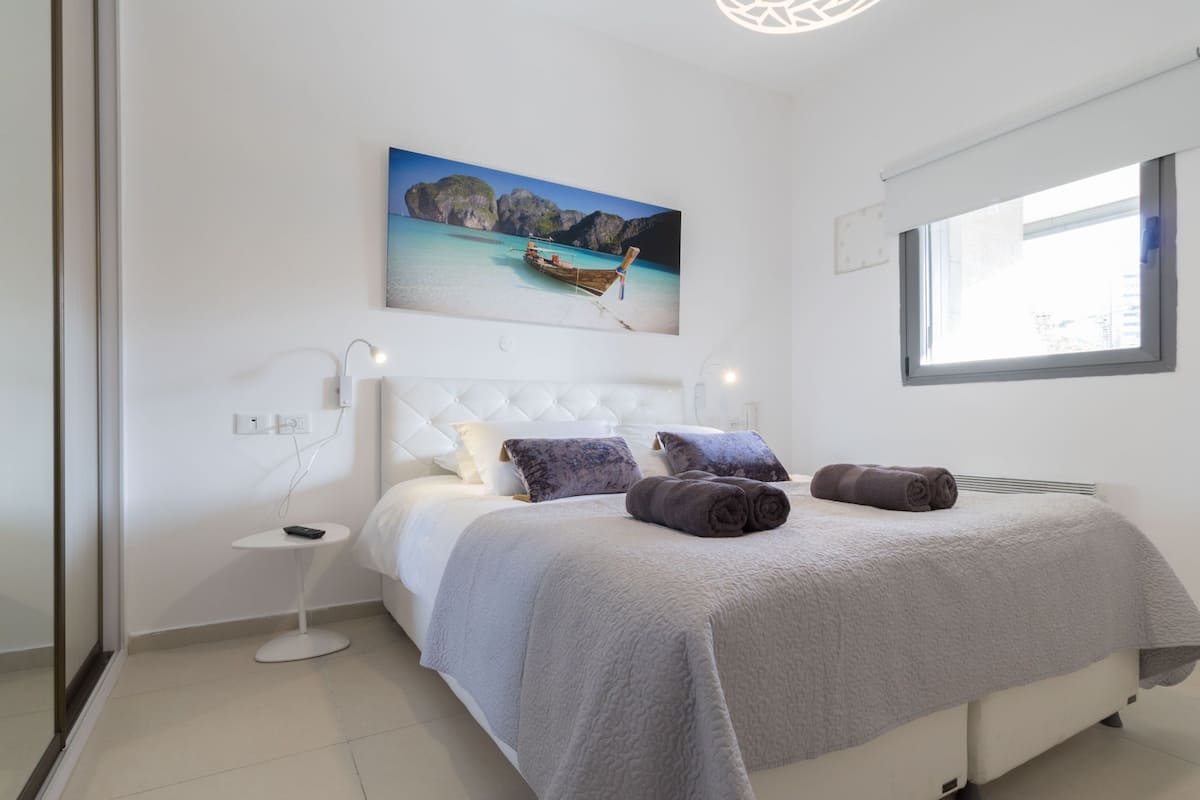 A modern bedroom with a bed, a nightstand, a painting of a beach scene, and a window.