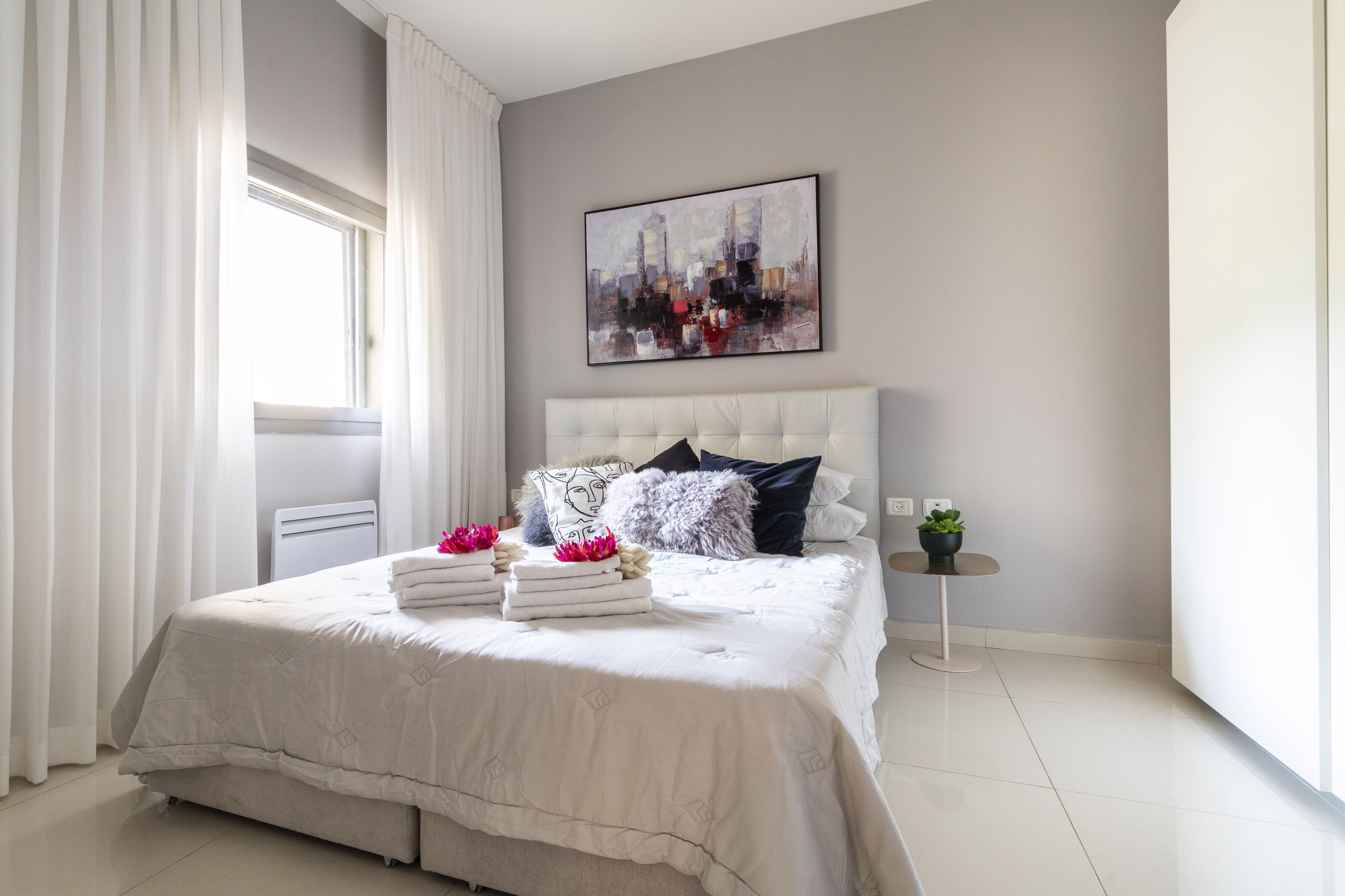 A modern bedroom with a large bed, white curtains, a cityscape painting, and a small round table with a plant.