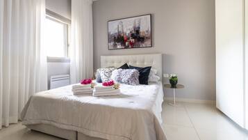 A modern bedroom with a large bed, white curtains, a cityscape painting, and a small round table with a plant.