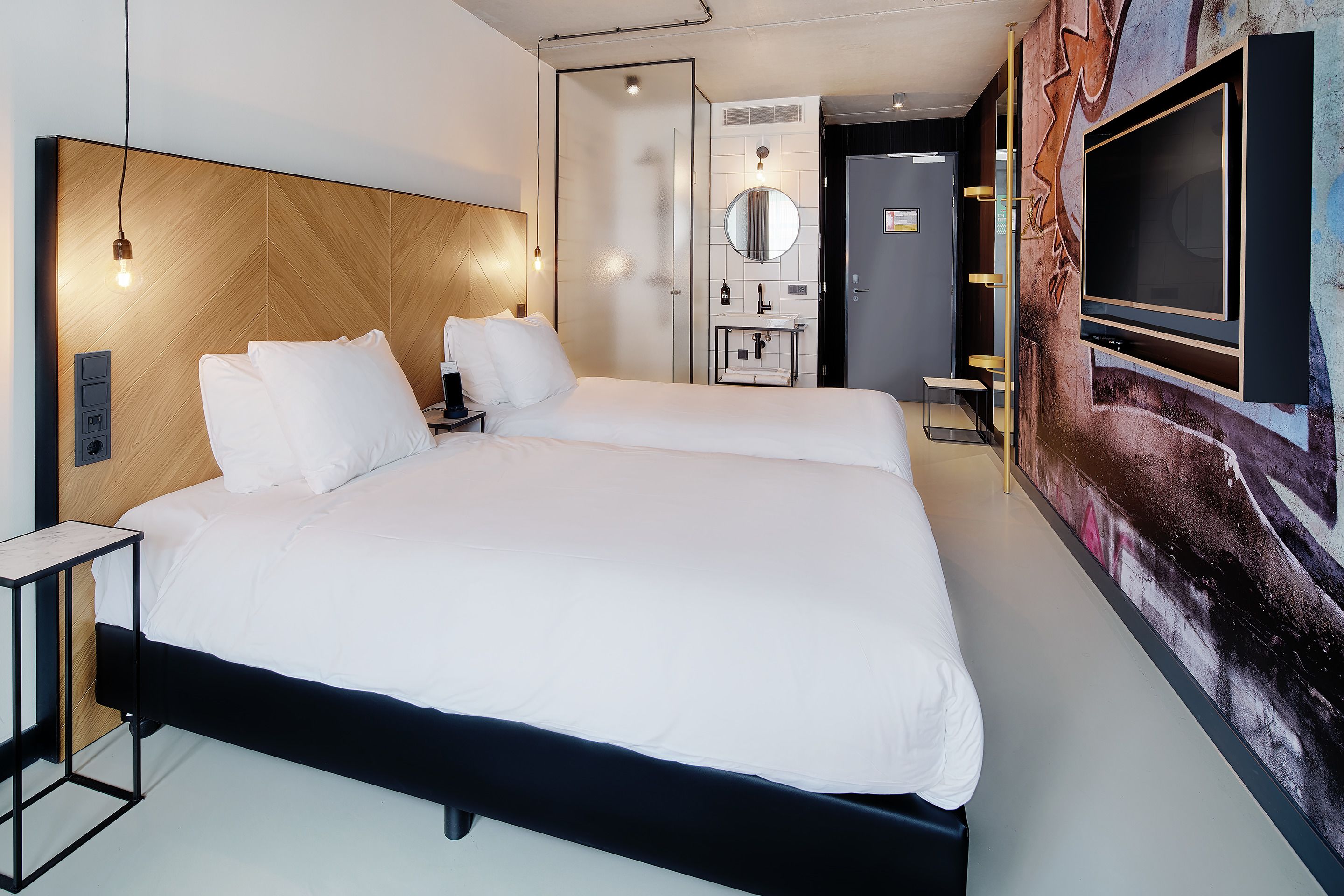Una habitación de hotel moderna con una cama grande, una mesita de noche, un televisor y un baño con espejo y lavabo.